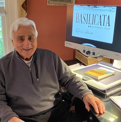 An older man with white hair is wearing a gray sweater and black pants. He is smiling as there is adaptive technology besides him on a desk. The screen reads "Basilicata".