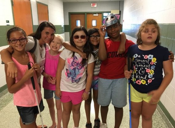 Group of children standing in hallway. One child wears sunglasses and 2 of the children hold mobility canes