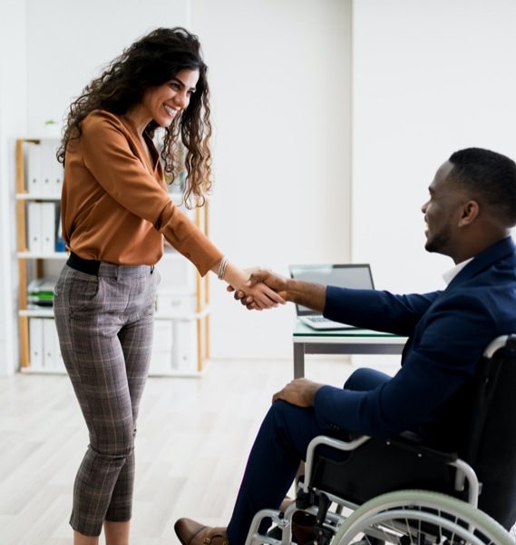 A smiling woman with dark curly hair shaking hands with a man with a short dark beard using a wheelchair