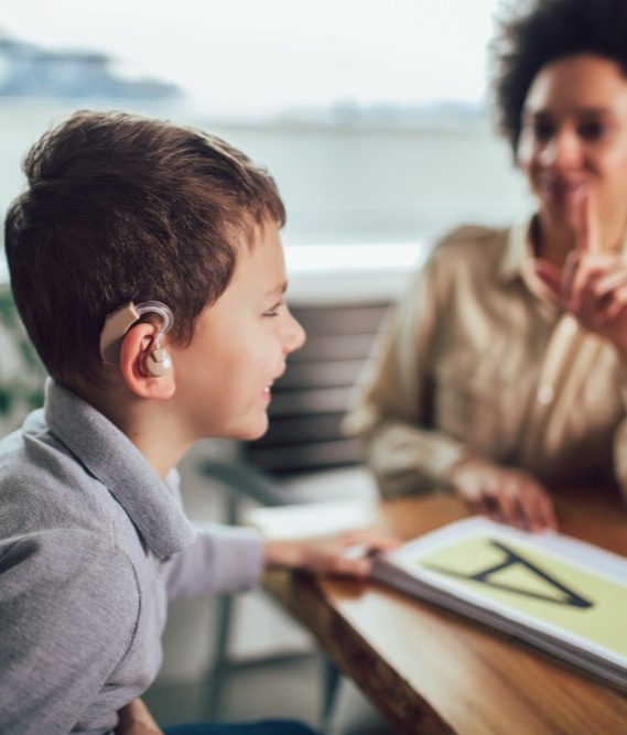 Child with hearing aid smiling at a desk next to an adult woman signing