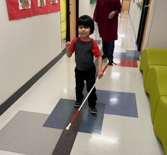A young boy walking down a hallway navigating with his white cane and an adult walking behind him.