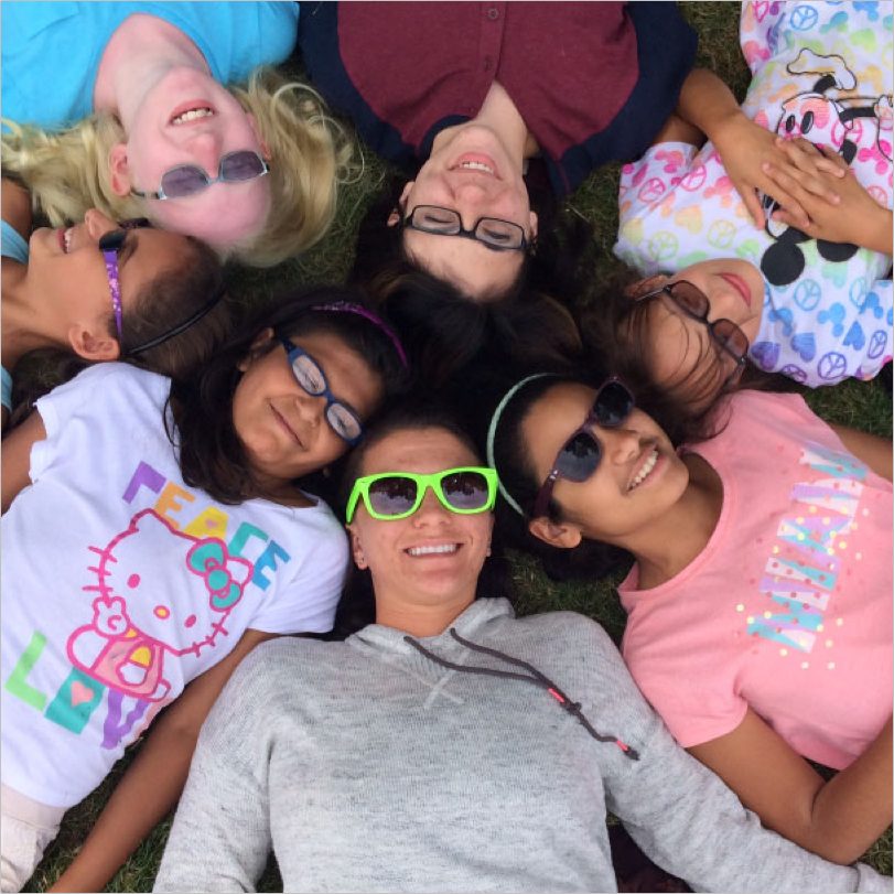 Seven kids laying on the grass with their heads in a circle.