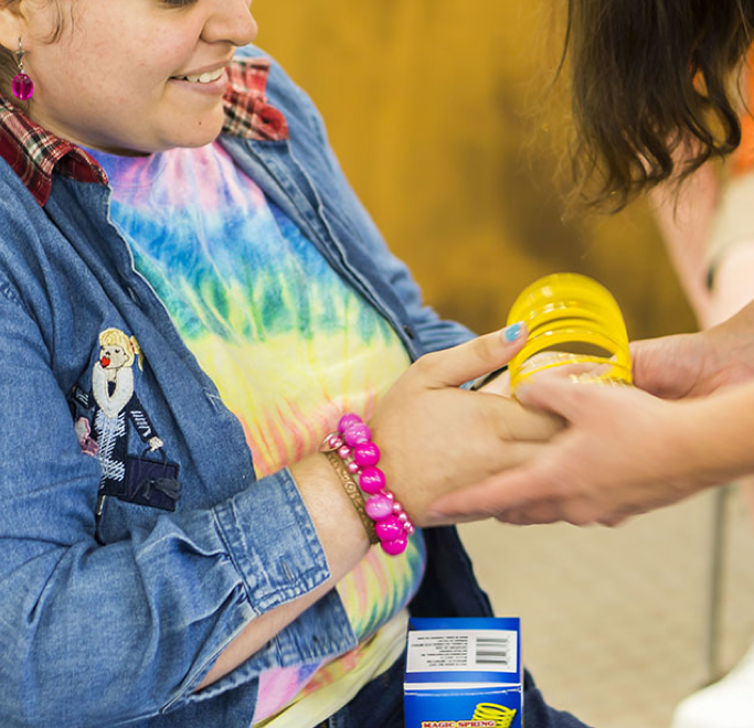 Two women holding a yellow object. One of the women wears a denim jacket and tie dye shirt 