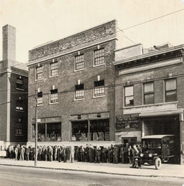 A large crowd of people and an old car in front of 2 buildings