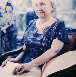 Helen Keller smiling, reading braille from a book, and wearing a fancy blue dress