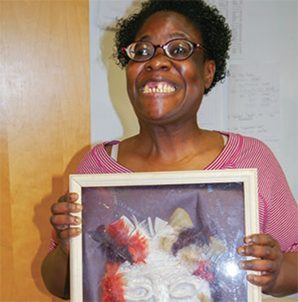 A woman wearing glasses and grinning widely holds a picture frame with art inside of it
