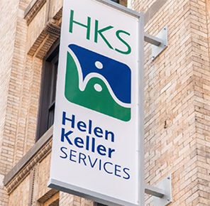 Wall signage that says “HKS Helen Keller Services” hanging perpendicular to a brick building