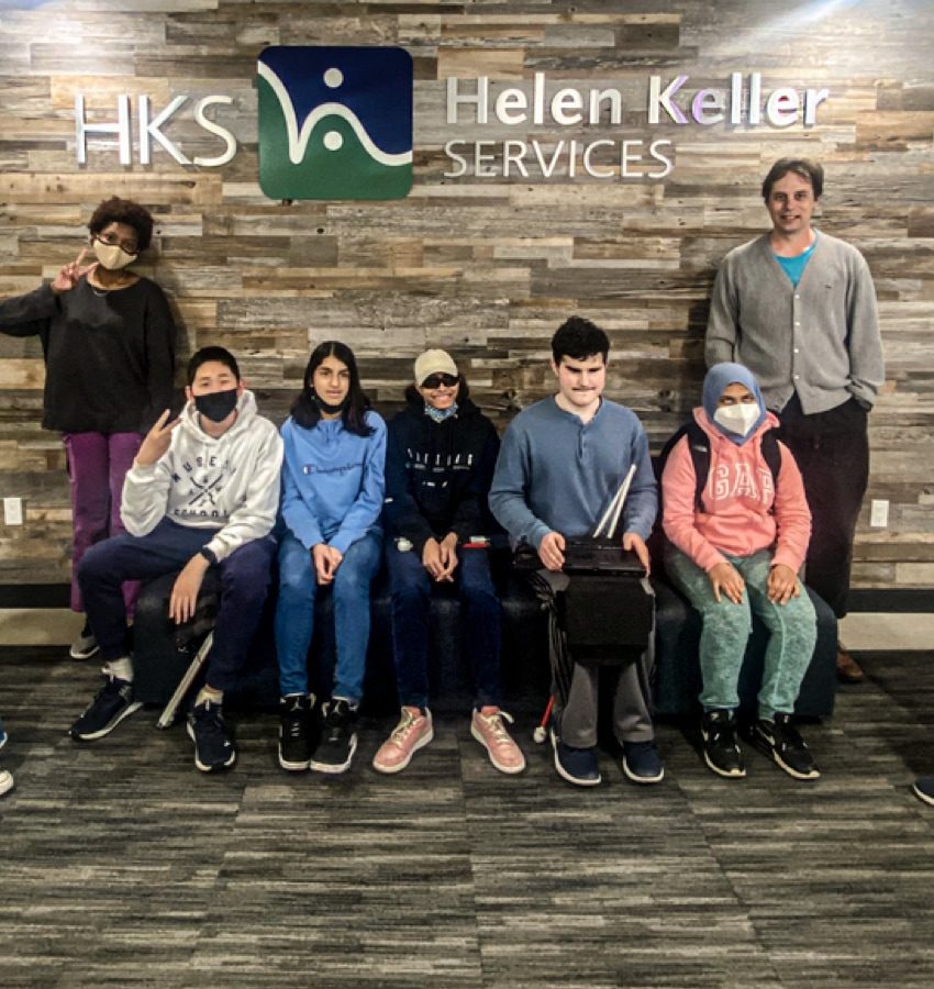 Group of boys and girls sitting and standing in front of the HKS Helen Keller Services sign that’s mounted on the wood-textured wall behind them 