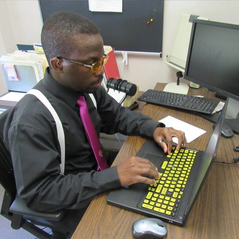 A man wears glasses, a hearing aid, and business attire and types on a laptop with large print keyboard