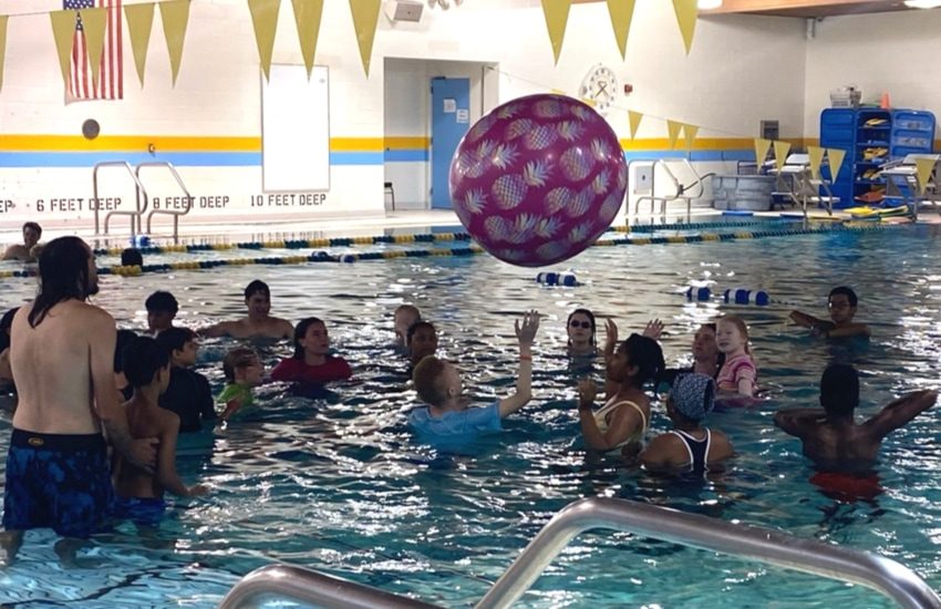 Group of campers in an indoor pool tossing around a large pink ball with pineapples on it 