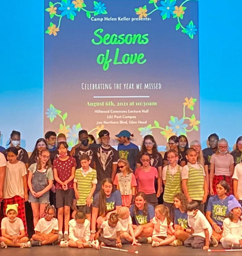 All campers and camp counselors on a stage with a screen behind them that says "Seasons of Love. Celebrating the year we missed."