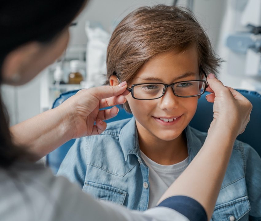 Boy with glasses sitting in office chair and smiling while woman adjusts his glasses