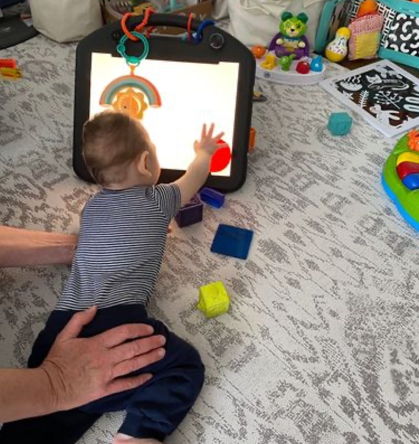 Baby reaches for propped-up tablet screen on floor while surrounded by baby toys and the guidance of adult hands on his bottom