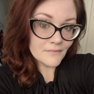 A Caucasian woman looking into the camera and smiling. She has long wavy copper hair, blue eyes, cat eye shaped glasses and is wearing a black shirt