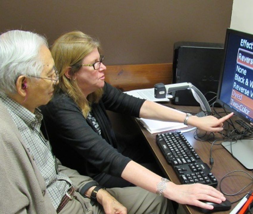 A woman and an older man with a hearing aid sit at a desk looking at a computer screen with large text on it. The woman is pointing at a computer screen with her left hand and holding the computer mouse with her right hand 