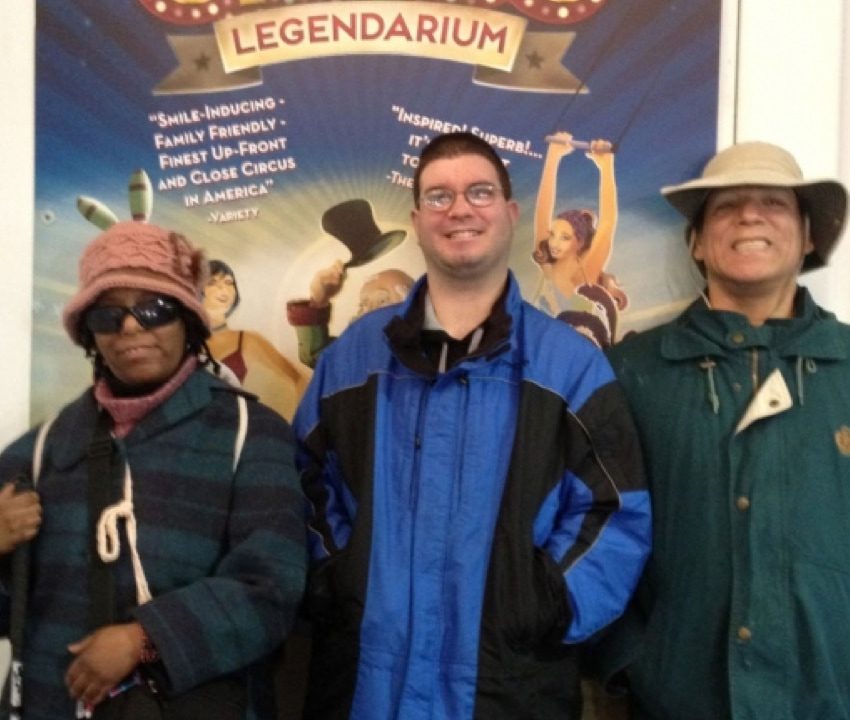 2 men and a woman smiling into the camera while standing in front of a large poster