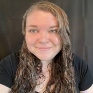 A Caucasian woman in front of a black backdrop staring at the camera and smiling. She has long curly brown hair, blue eyes, and is wearing a black shirt.