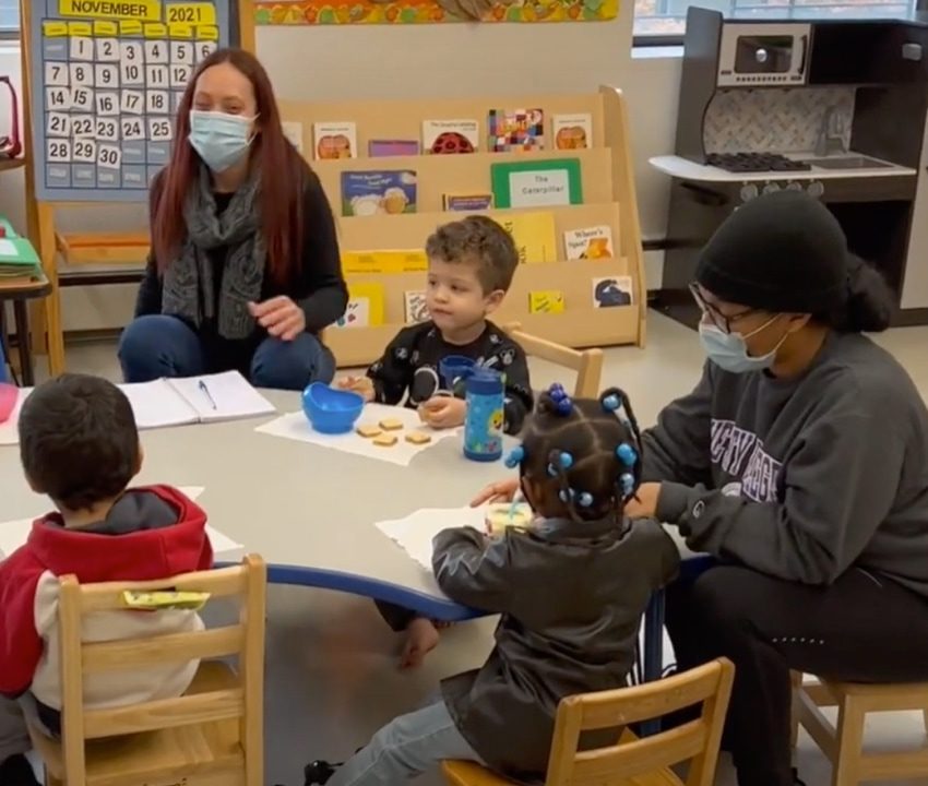 Group of 3 young children sitting at classroom table with snacks next to 2 adults wearing face masks