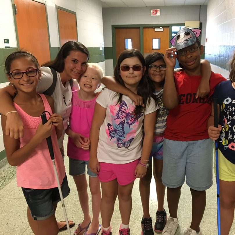 Group of children smiling in hallway. One child wears sunglasses and 2 of the children hold mobility canes