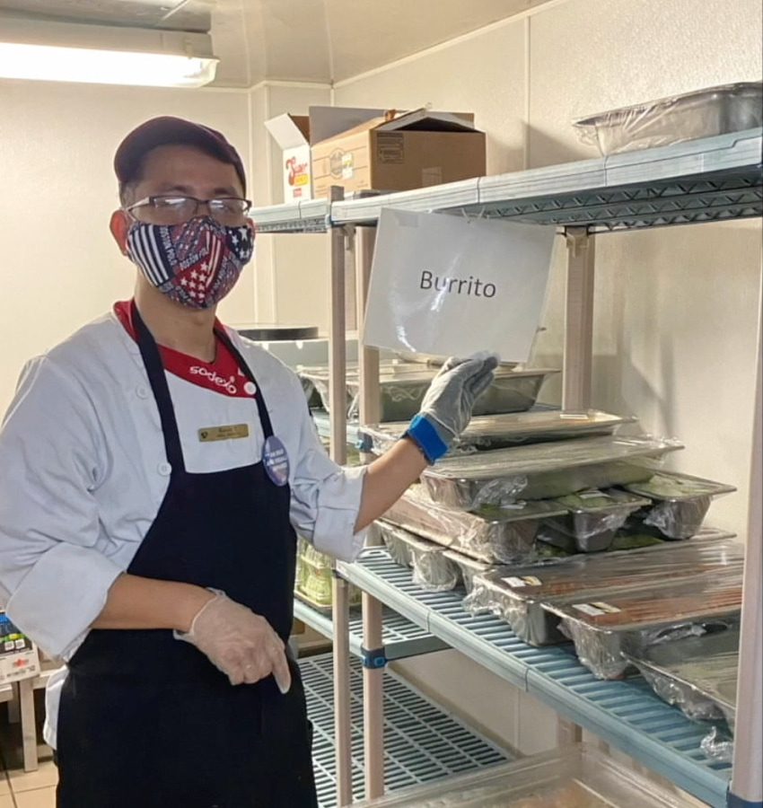 A man wears a Sodexo uniform, face mask, glasses, and apron and stands in a kitchen holding up a sign that says “Burrito” on it