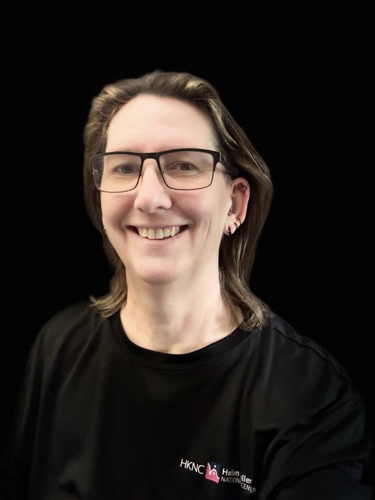 Head shot of Laurie, medium length brown hair with caramel highlights, wearing glasses and a black shirt with the Helen Keller National Center logo on the left side.