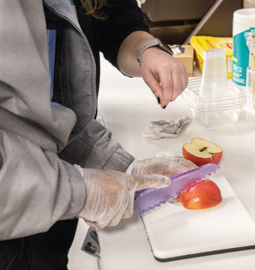 Person wears gloves and cuts an apple with a serrated purple knife while a person watches next to them