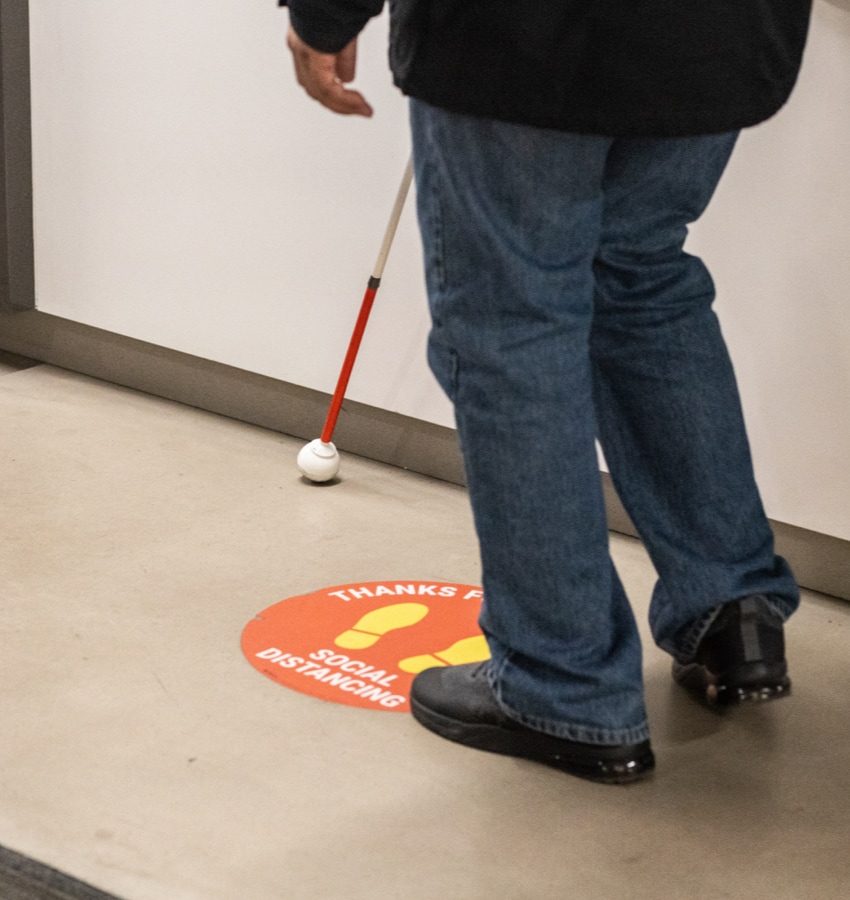 Person walking near hallway wall and using a white cane. The person stands on a decal says that “Thanks for social distancing” on a hallway floor