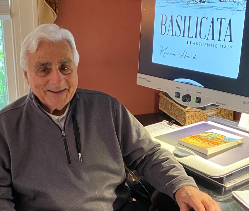 An older man smiling at the camera at a desk next to a computer screen that has the words “Basilicata, Authentic Italy, Karen Haid” on it in fancy typeface
