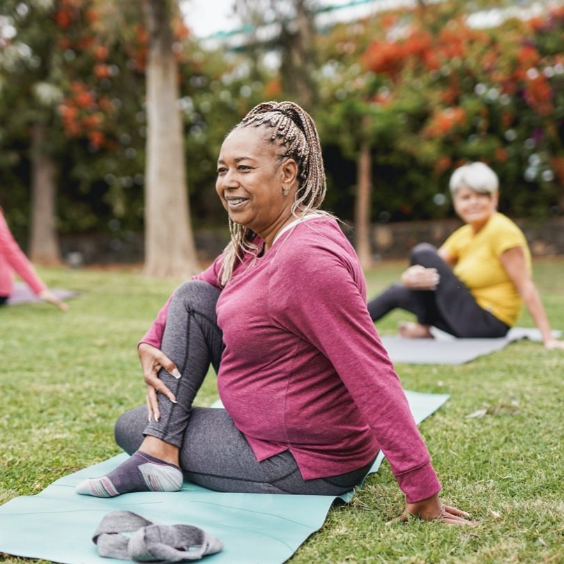 Woman with braids in a ponytail sitting on a yoga mat outside. Her right leg is bent over her left leg and she’s twisting her body to the left to engage in a back stretch