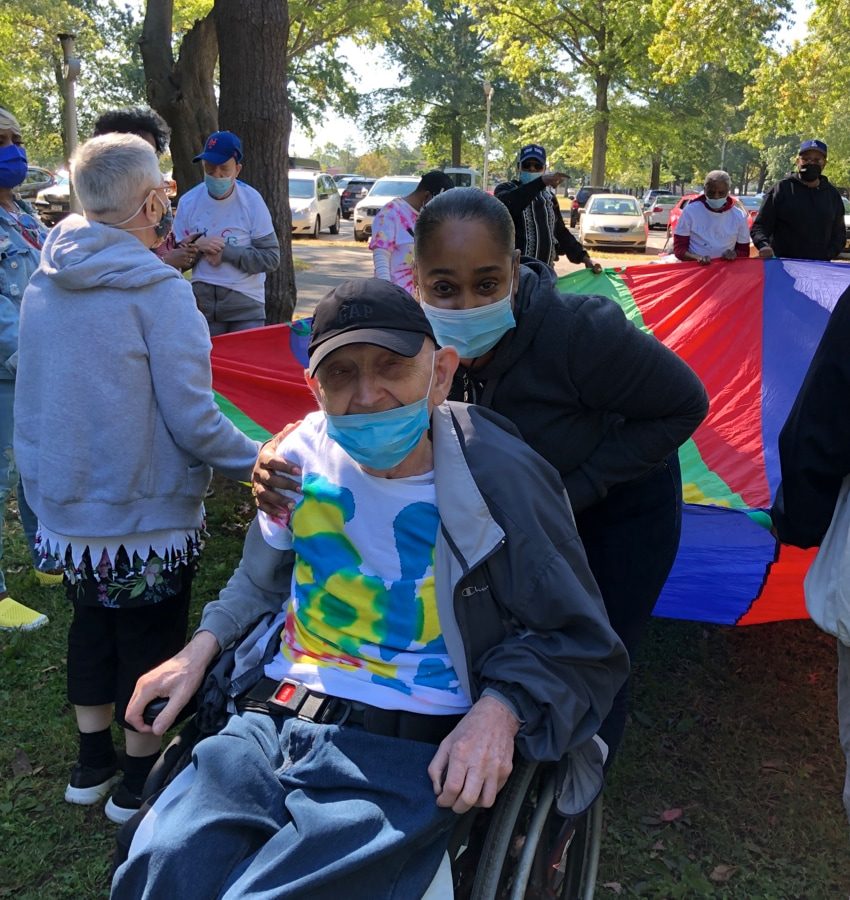 An older man using a wheelchair and woman smiling in front of a group of people playing with a rainbow parachute