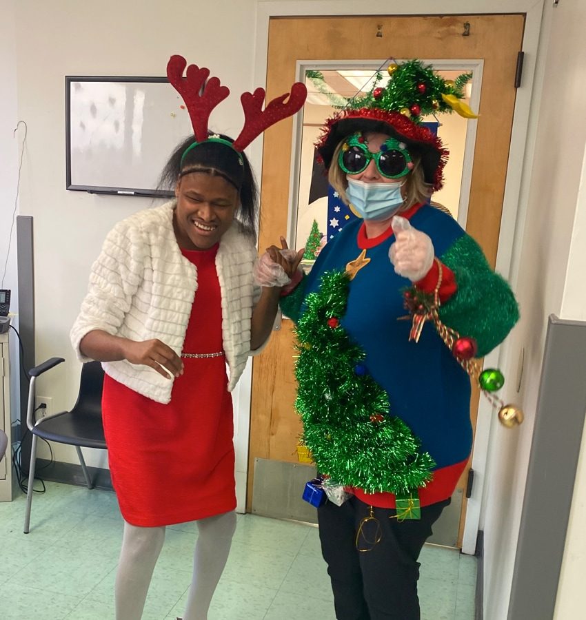 Two women dressed in colorful holiday clothing embellished with jingle bells, tinsel Christmas trees and reindeer antlers