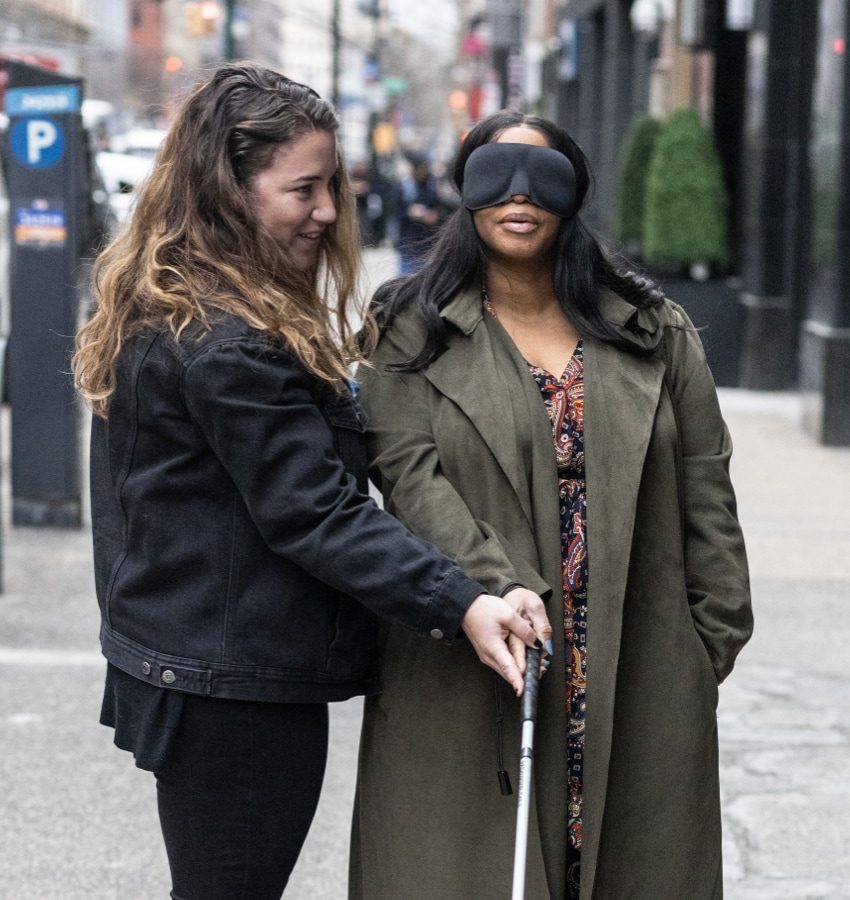 Two women stand on a sidewalk. One woman holds a white cane and wears a black mask over her eyes and the other woman has her hand on top of the other woman’s hand that’s on the cane