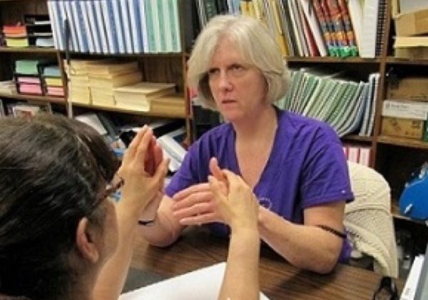 Two woman sit at a table and use tactile hand over hand sign 