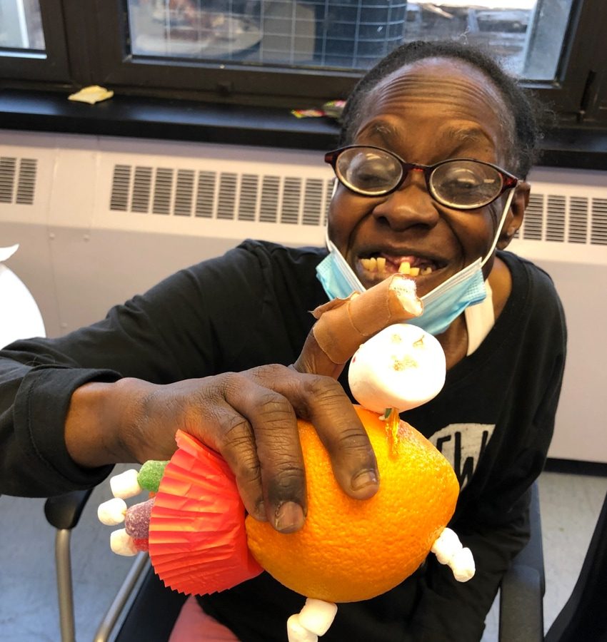 Middle-aged woman with glasses grinning while holding an art project made out of an orange, marshmallows, gum drops and a cupcake liner