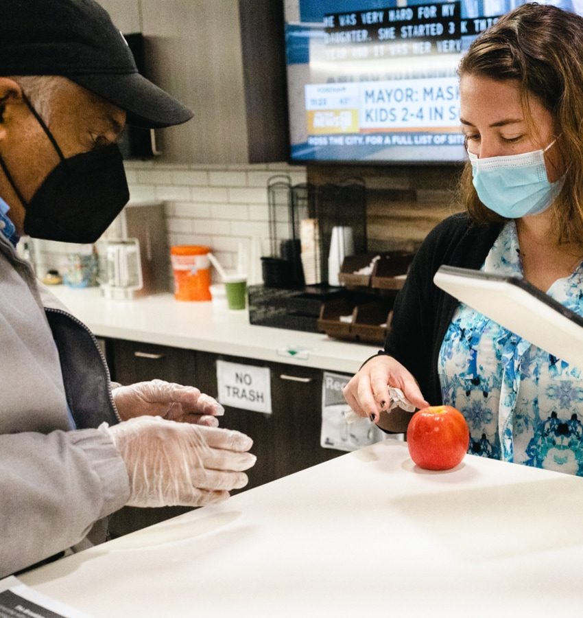 A man and a woman wearing face masks standing near a kitchen counter that has an apple on it