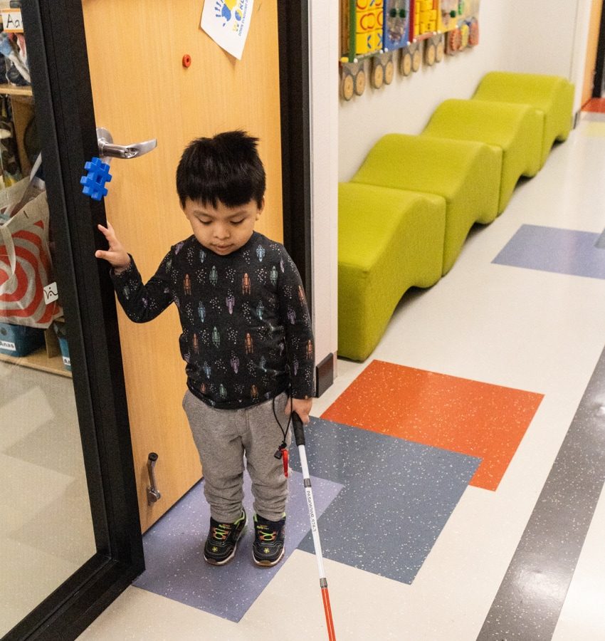 Young boy standing in a hallway holding white cane in left hand and touching door frame with right hand