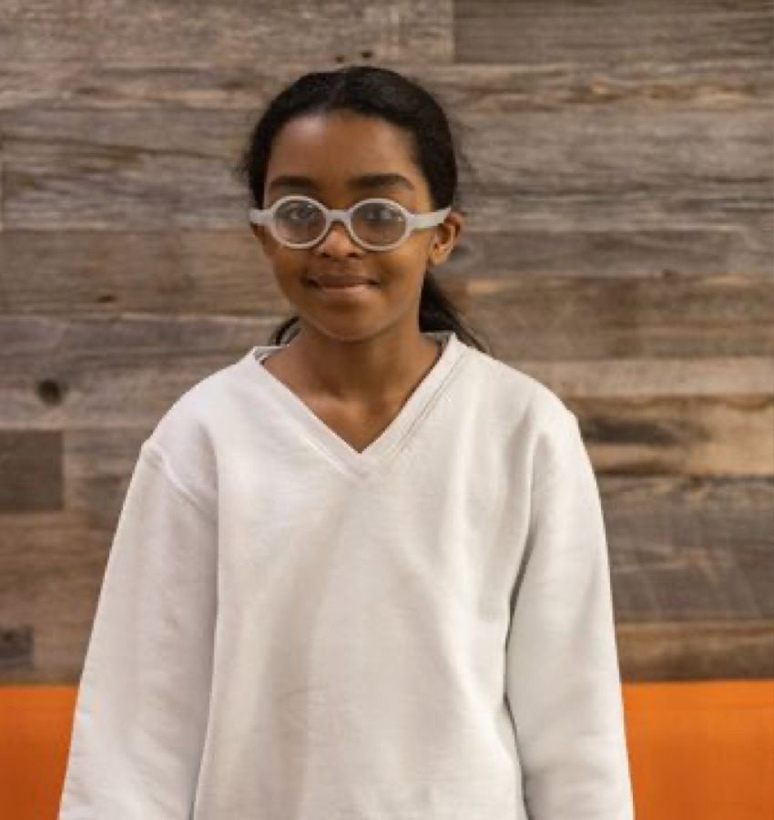 Girl wearing white sweatshirt and light grey glasses smiling into camera while standing in front of a wood-textured wall