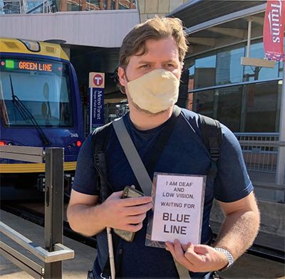Man standing in front of a bus terminal holding a white cane & device in his right hand and a sign in his left hand that says “I am deaf and low vision waiting for blue line.” 