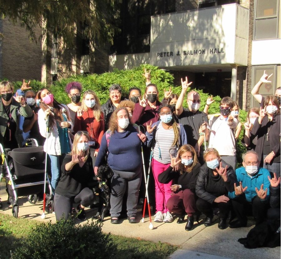 Large group of people standing outside, wearing face masks, and signing “I love you” in front of Peter & Salmon Hall at Helen Keller Services headquarters