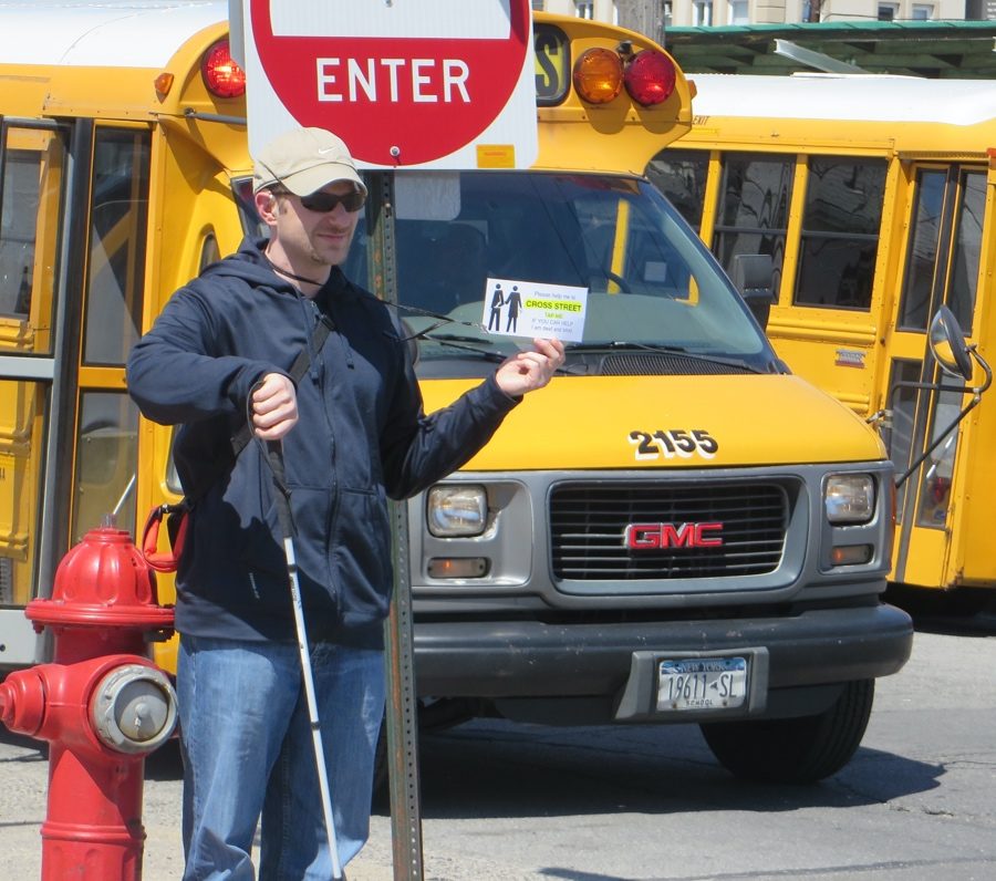 Man wearing sunglasses and a hat in front of a school bus holding a white cane in one hand and a street-crossing card in the other hand