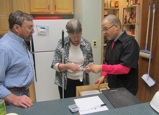 2 men and a woman standing in a kitchen. One of the men shows an adaptive device to the woman who holds a white cane