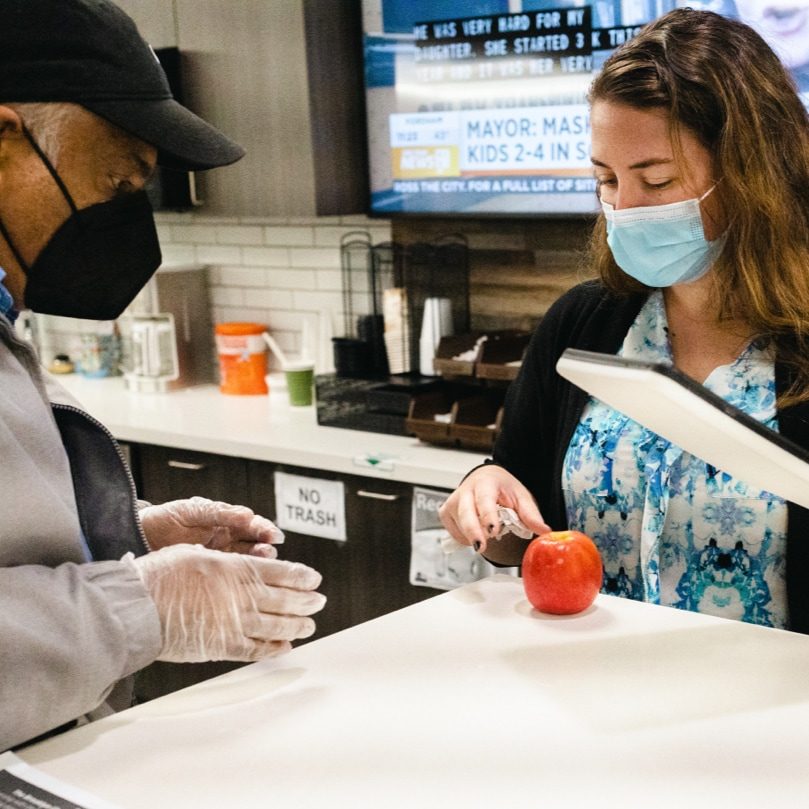 A man and a woman wearing face masks standing near a kitchen counter that has an apple on it