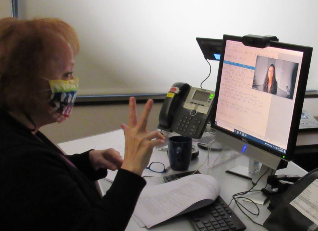 A woman signing to another woman on a computer that shows braille on the screen.