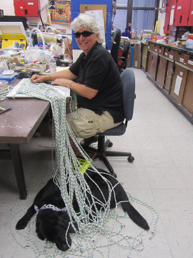 Person sitting in art studio wearing sunglasses and smiling into camera while working on macramé project. A black dog lays at her feet.