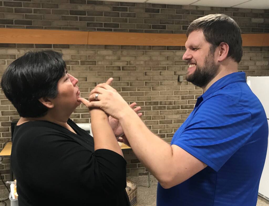 A woman dressed in a black shirt signing to a man wearing a blue shirt. Both are standing up in a room