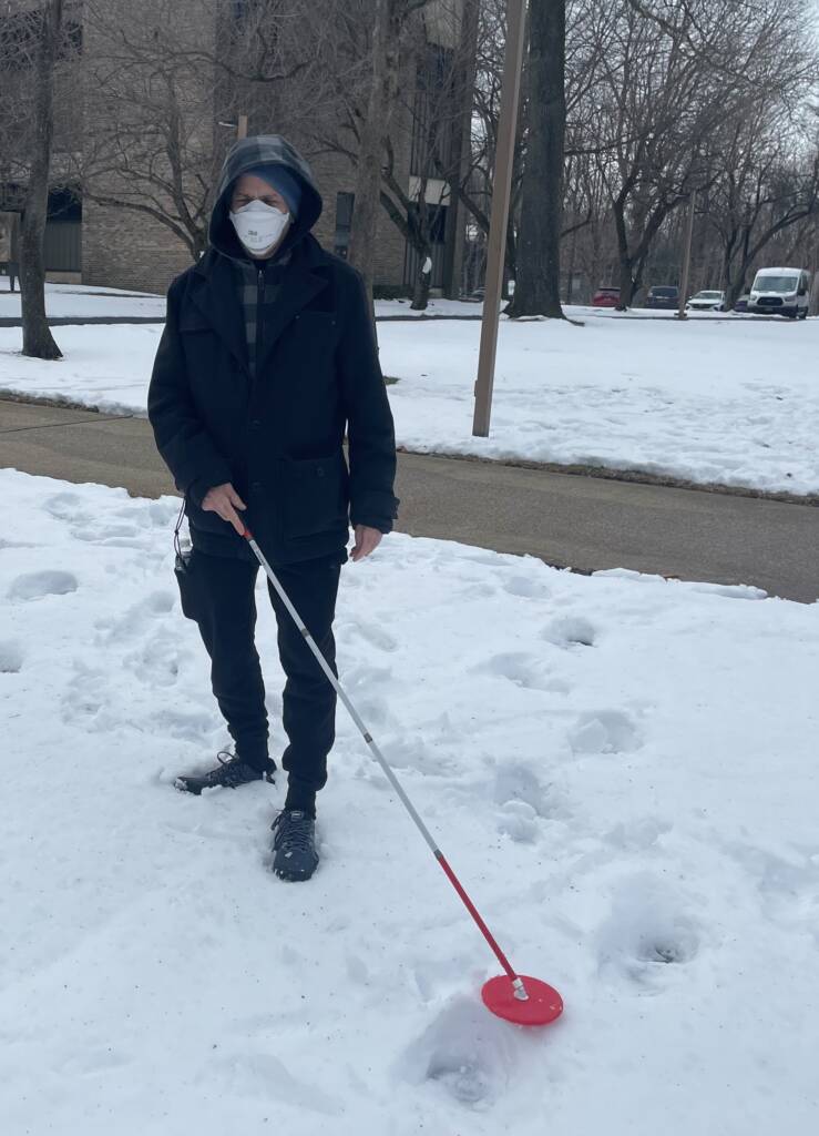 Man wearing face mask and jacket using white cane outside in the snow