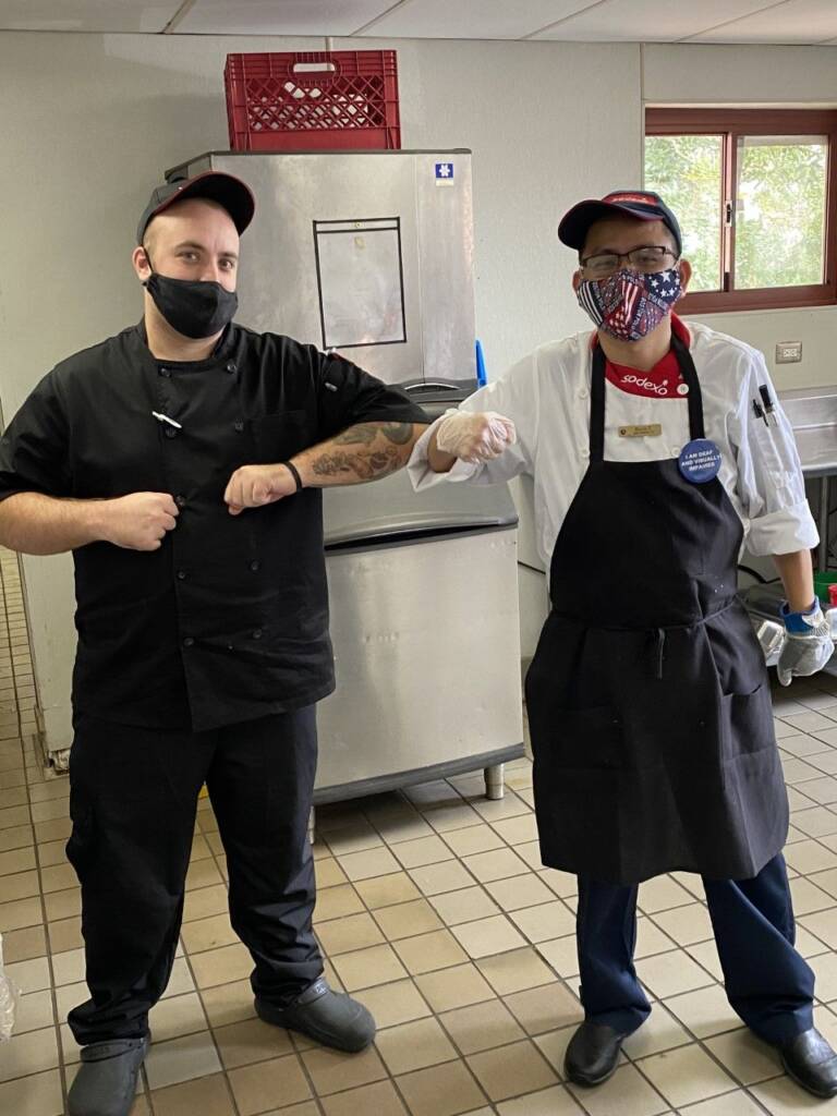 Two people wearing aprons, hats, and face masks standing in Sodexo kitchen and touching elbows