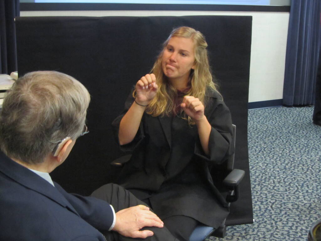 A woman dressed in black signing to a man wearing a hearing aid and suit jacket. Both people are sitting in a room together