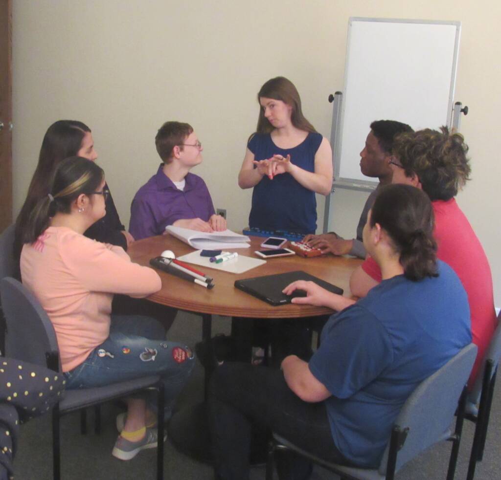 A group of young men and women seated around a small circular table and looking at a woman standing near them