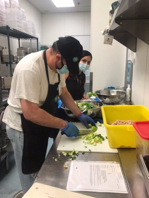 Person wearing face mask cutting vegetables in a restaurant-style kitchen under supervision of staff member